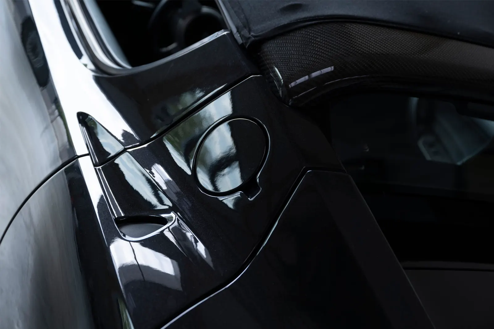 Detail an der Flanke eines schwarzen Tesla Roadster: die runde Klappe des Ladeanschlusses und der Übergang zum Windlauf, im Lack spiegelt sich die Halle.