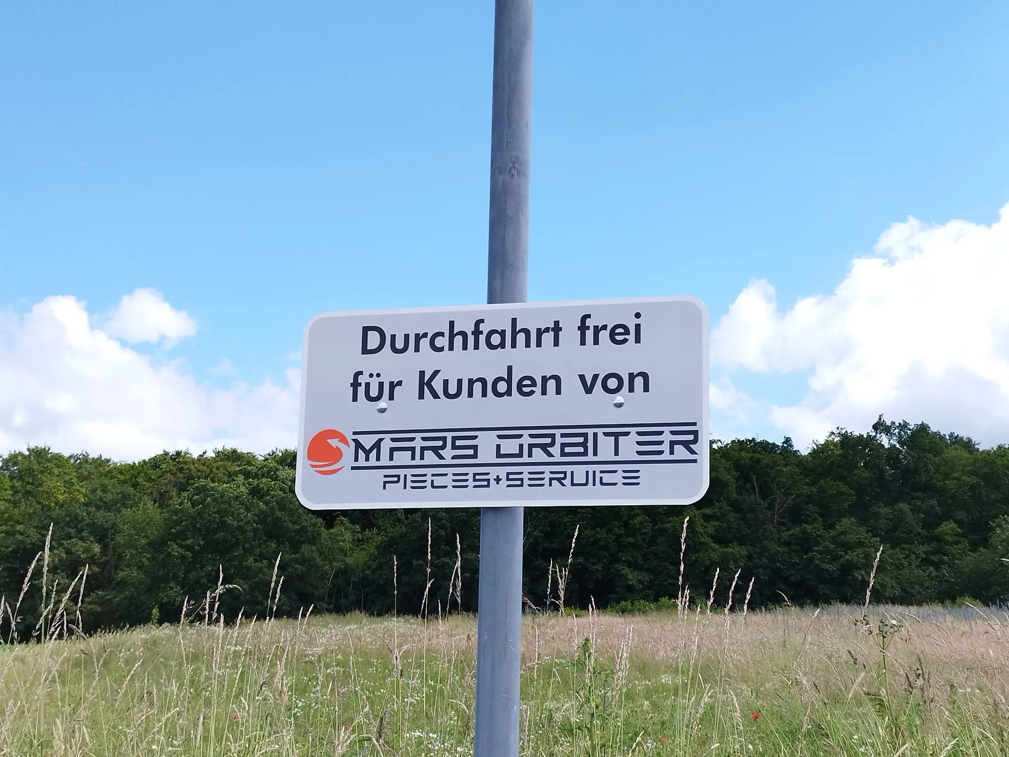 Hinweisschild „Durchfahrt frei für Kunden von Mars Orbiter" an einem Mast auf dem Weg zur Werkstatt.